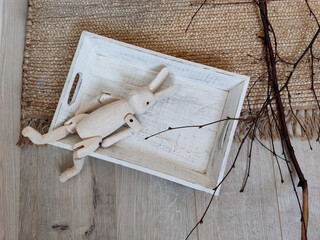 Overhead, flat lay shot of a rustic scene featuring a light-colored wooden rabbit toy with jointed limbs resting inside a distressed white wooden tray, set on a jute rug 