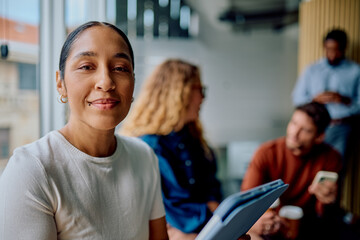Confident businesswoman smiling at camera, holding a notebook, with diverse colleagues collaborating in a modern office background
