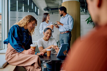 Group of diverse coworkers having a casual office break, using digital tablet and smartphones, drinking coffee and laughing