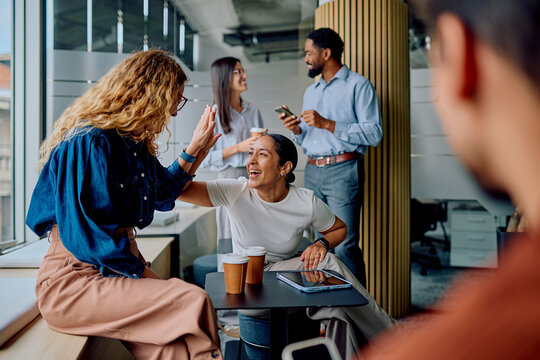 Diverse business colleagues high-fiving, smiling, and discussing in a modern office, celebrating collaboration and achievement during a coffee break