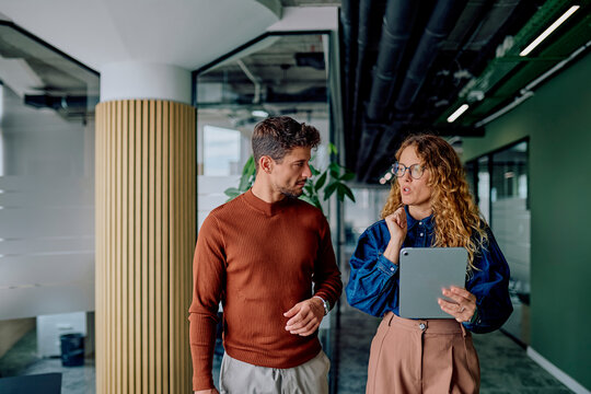 Two business colleagues walking and discussing work in a modern office corridor, focused on a tablet