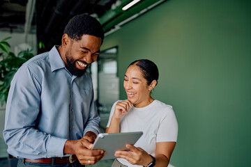 Diverse business colleagues standing in an office, smiling and laughing while looking at a digital tablet screen together