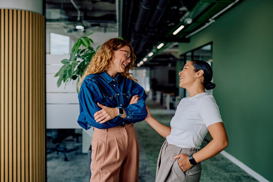 Two diverse women colleagues laughing and talking in a modern office hallway, showcasing teamwork and professional friendship - Powered by Adobe