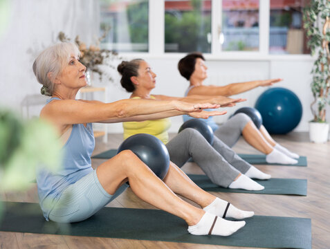 Calm mature woman doing pilatess sitting on black mat with soft ball between legs during workout session