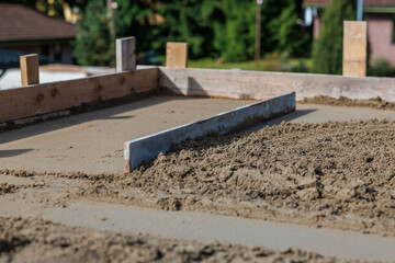 A construction site featuring a concrete slab framed by wooden boards. A metal screed tool is visible, with blurred structures and vegetation nearby.