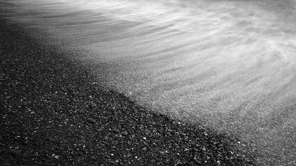 Black and white beach with a wave crashing into the shore. The water is foamy and the sand is dark, creating a moody and dramatic atmosphere