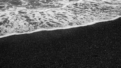 Black and white beach with a wave crashing into the shore. The water is foamy and the sand is dark, creating a moody and dramatic atmosphere.