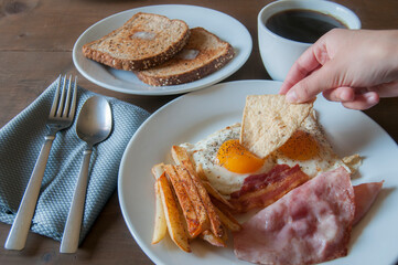 Fried eggs with ham and French fries on a white plate served as breakfast with black coffee and toast with butter on a wooden table with a 45-degree viewing angle, tortilla chip