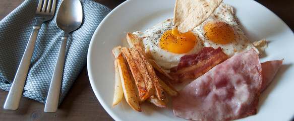 Fried eggs with ham and French fries on a white plate served as breakfast with black coffee and toast with butter on a wooden table with a 45-degree viewing angle, panoramic