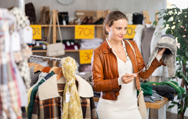 European woman customer examine and choose fur hat in store, and buyer consider some items. Concept of seasonal wardrobe shopping. Client enjoys shopping for stylish clothes