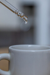 Detalle macro de una pipeta transparente dejando caer una gota sobre una taza de cerámica blanca,...