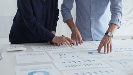 Business colleagues are examining financial data, pointing at charts and graphs spread out on a table during a collaborative analysis session in a modern office. - Powered by Adobe