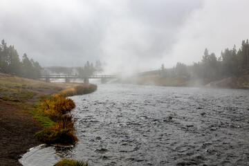 Serene river flows under a wooden bridge amidst misty landscape, evoking tranquility and the beauty...