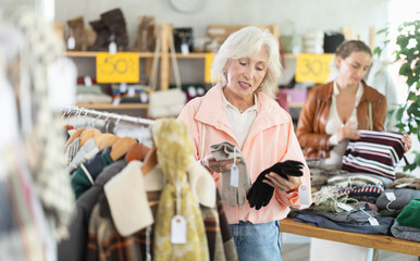 Mature woman with gray hair standing in store and choosing gloves in front of female shopper looking at counter with sweaters