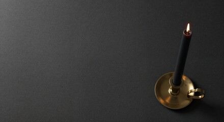 A top-down view of a single elegant black candle with a lit flame in a shiny brass candlestick on a dark gray textured background, offering generous negative space.