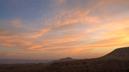 Warm hues fill the sky as the sun sets behind distant mountains in a serene desert landscape. The soft clouds enhance the tranquil scene, highlighting nature's beauty at twilight. Guadalupe National 