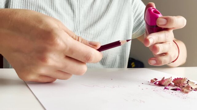 Close up video of woman sharpening pencil with pink sharpener over white paper on desk. Stationery use, school preparation, office supplies, drawing tool, creative process.