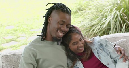 Diverse couple exchanging smiles leaning heads and wrapping arms on patio sofa for affection - Powered by Adobe