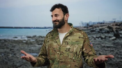Hispanic man with beard in military uniform standing on rocky beach with sea backdrop, looking confident and expressive, blending military presence with tranquil coastal scenery. - Powered by Adobe