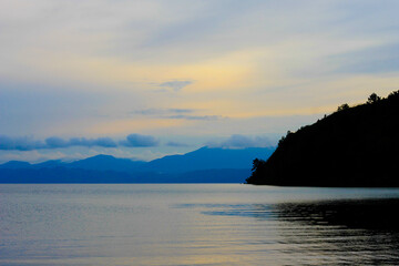 Foto lanskap alam yang menampilkan keindahan danau dengan air biru jernih, dikelilingi perbukitan hijau yang rimbun. Di tepi danau terlihat rumah-rumah sederhana serta pepohonan tropis yang asri, menc