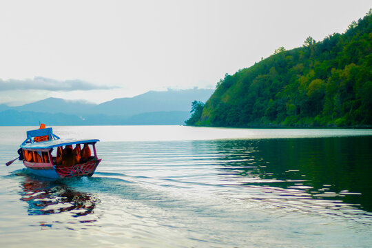 Foto perahu kayu tradisional dengan atap biru membawa penumpang menyusuri danau yang tenang. Air memantulkan bayangan perahu dan bukit hijau di sekitarnya, menciptakan suasana damai dan alami. Pemanda