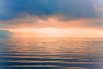 Fototapeta premium Foto pemandangan laut saat matahari terbenam dengan suasana hangat dan tenang. Cahaya matahari oranye keemasan menembus awan gelap dan memantul di atas permukaan air yang beriak lembut, menciptakan nu