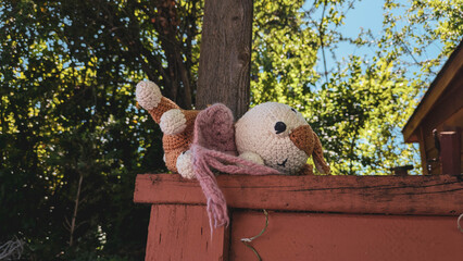 Close up of a cute crocheted stuffed animal abandoned on a fence with a tree and branches in the background. 