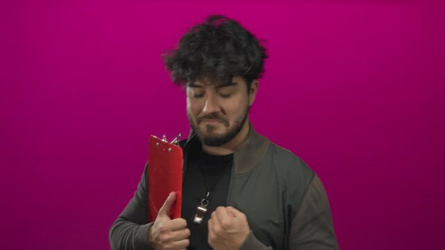 Young hispanic man with beard holding clipboard against pink background wearing jacket and whistle looking determined and thoughtful