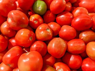 A large, full-frame shot of a pile of vibrant red tomatoes. The fruits vary in shape and size, with one prominent green tomato visible in the top center, creating a fresh and natural market display.
