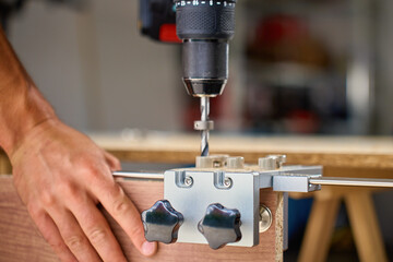 Close-up of carpenter drilling hole in wooden panel using drill and metal jig tool for precision. Man working in home workshop. Concept of woodworking, furniture making and DIY home projects