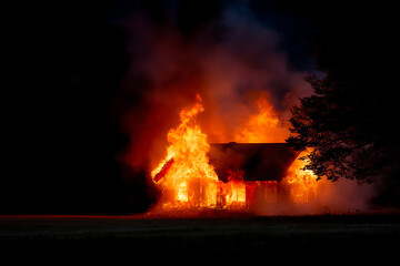 A house engulfed in intense flames at night, with thick smoke billowing into the dark sky and a tree silhouetted nearby