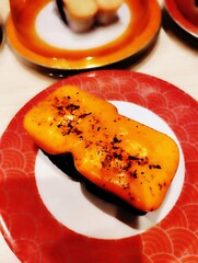 A Close-up photo of a sushi dish served on a white plate with a red patterned rim. The sushi is topped with a generous layer of melted orange cheese with red seasoning on top.