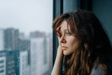 Young woman stares out rain-speckled window at blurred cityscape, inwardly pensive and solitary,...