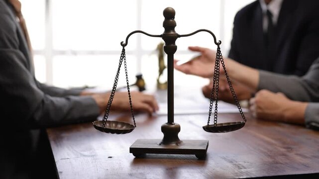Balance scale resting on wooden table with legal professionals discussing contract details during professional meeting inside law office.