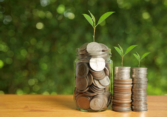 Coins in glass jar and stacked piles with small green plants growing, symbolizing savings, financial growth, investment, wealth management, sustainability, and long-term economic success.