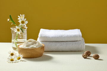 Glass jar with fresh chamomile, towel and bowl of shea butter on bright yellow backdrop, styled as spa-inspired organic cosmetic scene.