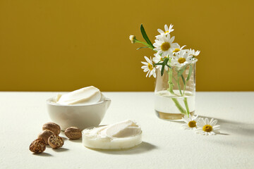 Simple still life with shea butter cubes, scattered nuts and chamomile flowers on clean white surface, designed for cosmetic ingredient visuals.