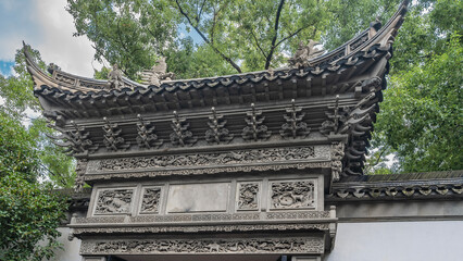 Ancient Chinese architecture. A fragment of a building with curved roof edges, carvings on cornices. Green branches of trees against a background of blue sky and clouds. China. Yu Garden. Yuyuan