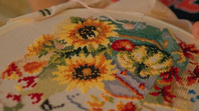 Close-up of bright cross-stitch with sunflowers, red apple and various colorful patterns. A woman's hand with a needle makes new crosses