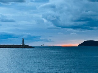 The lighthouse of Bar, Montenegro, standing on the Adriatic coast during a dramatic sunset, with storm clouds gathering over the sea and waves crashing against the rocky shore