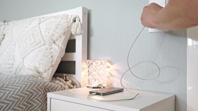 Close-up of a hand plugging a smartphone charger into a wall socket next to a bed with a decorative lamp, showcasing charging and uncharging actions in a domestic setting