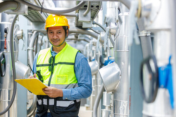 Industrial engineer in safety vest and hard hat holding checklist and walkie-talkie while inspecting pipeline systems in factory environment, ensuring safety standards and equipment performance.