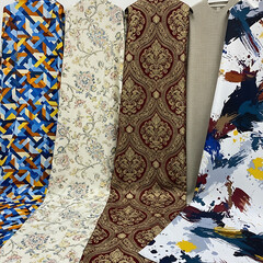 A diverse collection of folded fabrics presents an array of textures and patterns, including floral, tweed, velvet, lace, and stripes, creating a rich and varied textile display