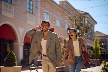 Young carefree couple holding hands and running while shopping in city.