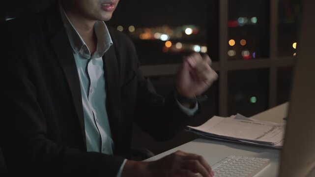 Night office worker in suit using desktop computer with serious expression, hardworking Asian business professional in dark interior with city lights, stress, focus and corporate lifestyle.