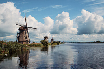 Fototapeta premium A scenic view of traditional windmills lining a waterway under a dramatic blue sky filled with white clouds