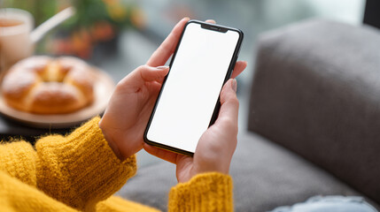 A person wearing a warm yellow sweater sits indoors and holds a modern smartphone with a blank white screen for mockup purposes