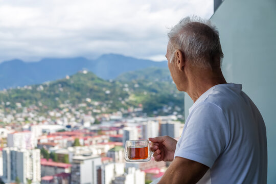An elderly man drinking tea looks at the green mountain city, standing on the balcony of a skyscraper. The idea of moving to a warm country for retirement