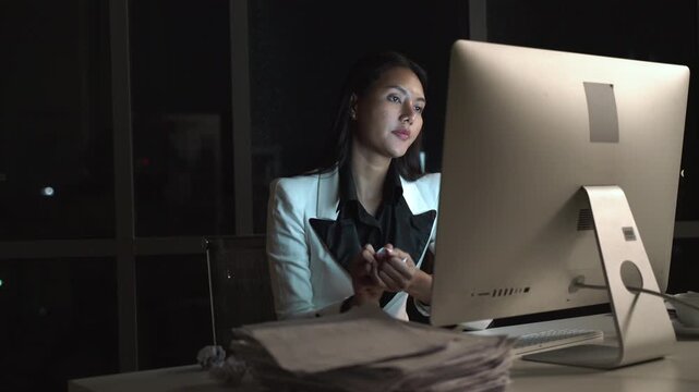 Hardworking businesswoman working late at night in a modern office, focused on desktop computer screen, determined female executive facing deadline under dim lighting with stack of documents.