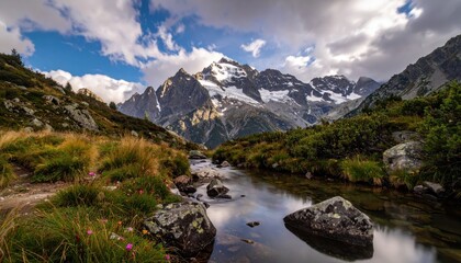 Fototapeta premium Dramatic landscape photograph captures a tranquil stream meandering through a vibrant, grassy meadow, with imposing, snow-capped mountain peaks dominating the background under a dynamic, partly...